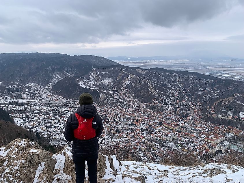 A female hiker in winter hiking gear stands atop a snowy mountain, looking down on a quaint mountain town. 