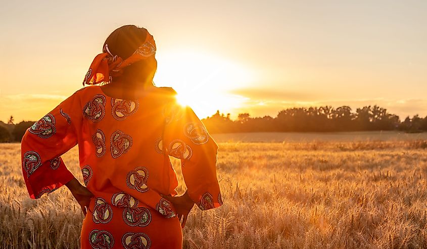 African woman in traditional clothes standing with her hands on her hips in field of barley or wheat crops at sunset or sunrise