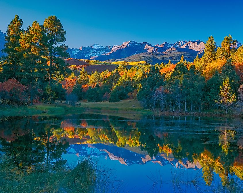 A pond surrounded by fall color reflects the trees and the Sneffels Range mountains in Ridgway, Colorado