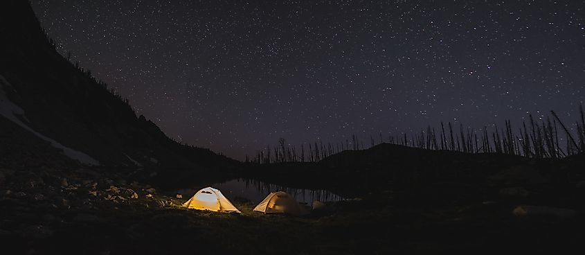 Lit tent at night in the Sawtooth Mountains of Idaho with stars in the sky above next to an alpine lake.