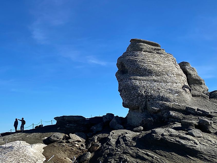 A large boulder with a humanoid face watches a couple of hikers.