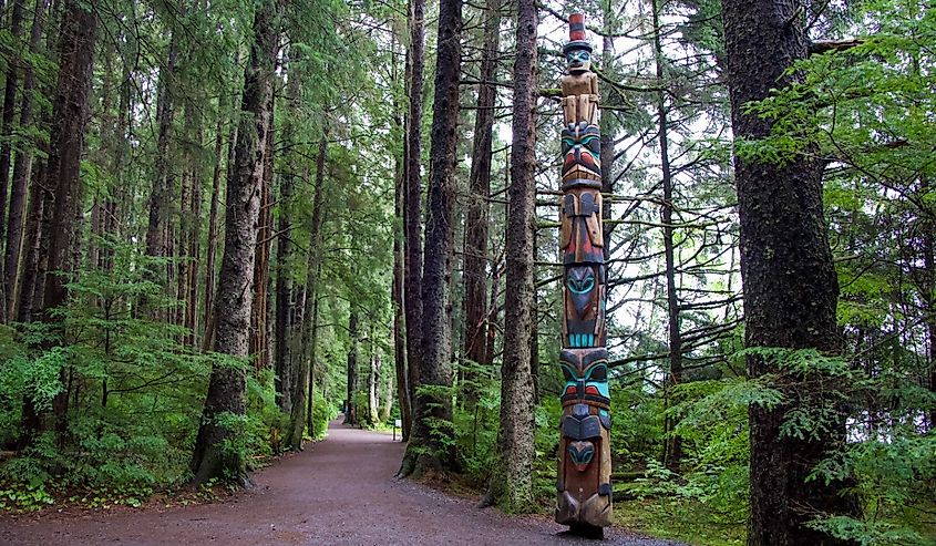 Alaska totem pole on walkway pathway in forest with tall pine trees and green trees in woods, Sitka, Alaska.