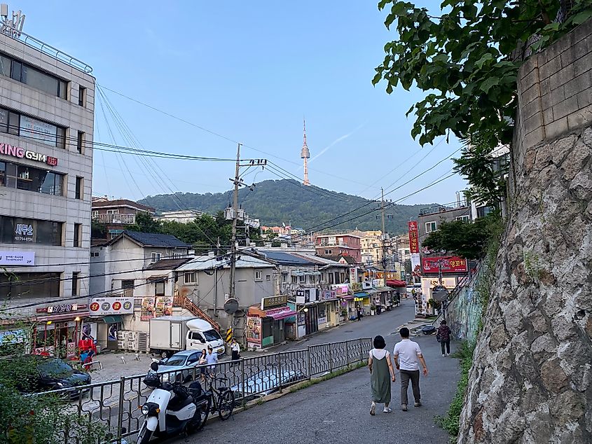 A couple walks down a ramp, beside a stone wall, and toward the restaurants of a small neighborhood in Seoul.