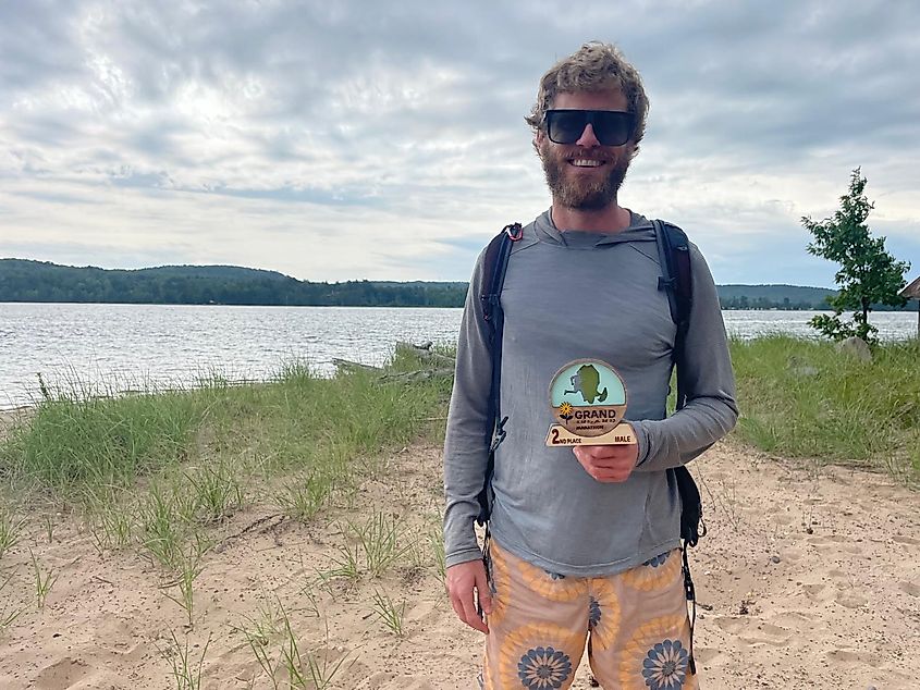 A man poses on a wild island beach with a wooden trophy stating 2nd Place Male in the Grand Island Marathon.