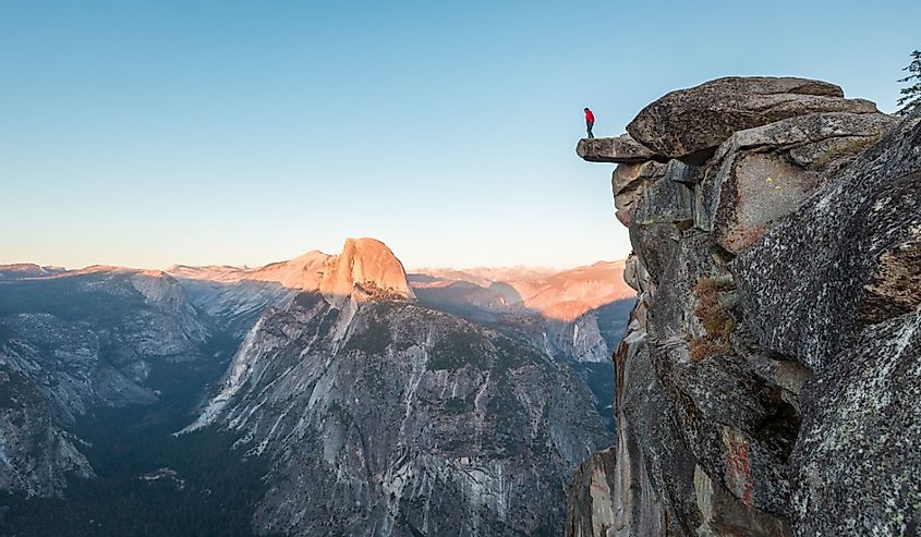 A fearless hiker is standing on an overhanging rock enjoying the view towards famous Half Dome at Glacier Point overlook in beautiful evening twilight, Yosemite National Park, California