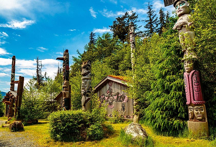 A serene outdoor scene with tall, intricately carved totem poles surrounded by lush greenery. A wooden building adorned with native art is in the background under a bright blue sky.
