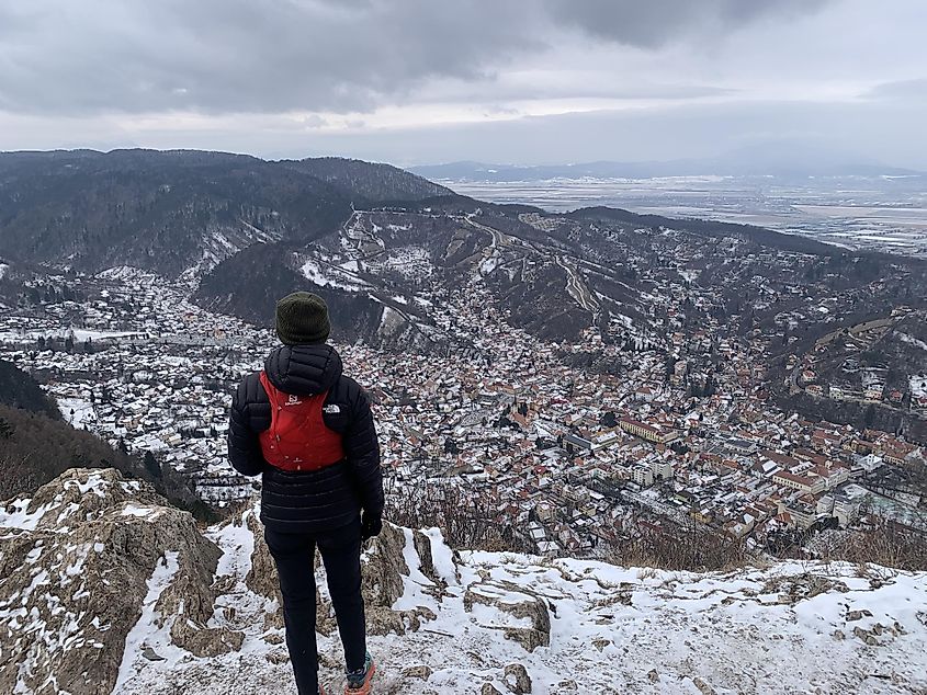 A female hiker stands atop a mountain, looking down on the snowy Old Town of Brașov, Romania.