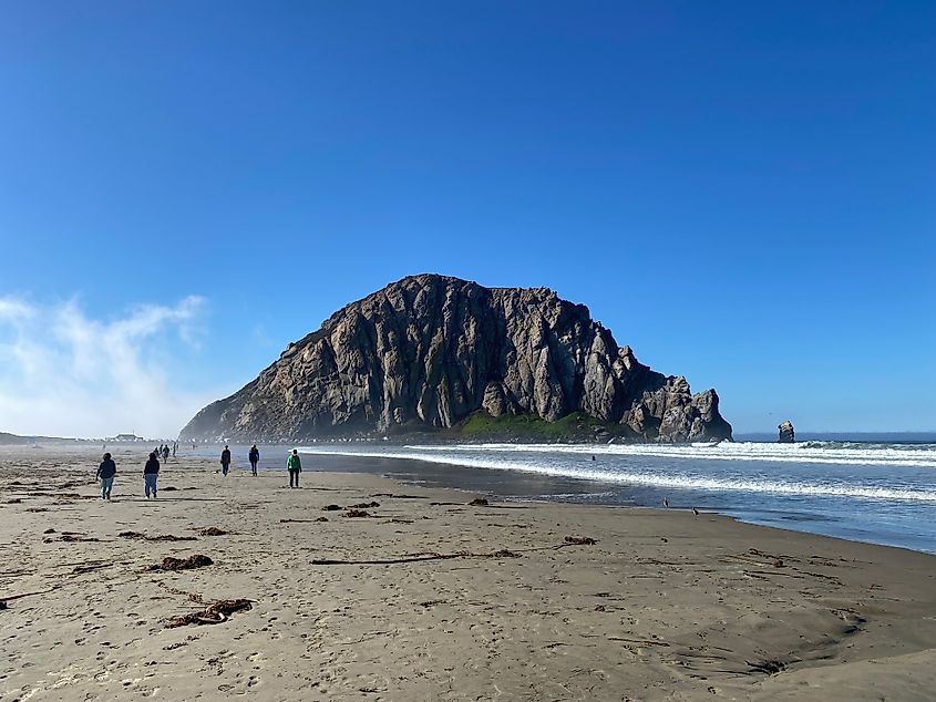 People walking toward a large volcanic rock monolith that stands at the end of a large ocean beach.