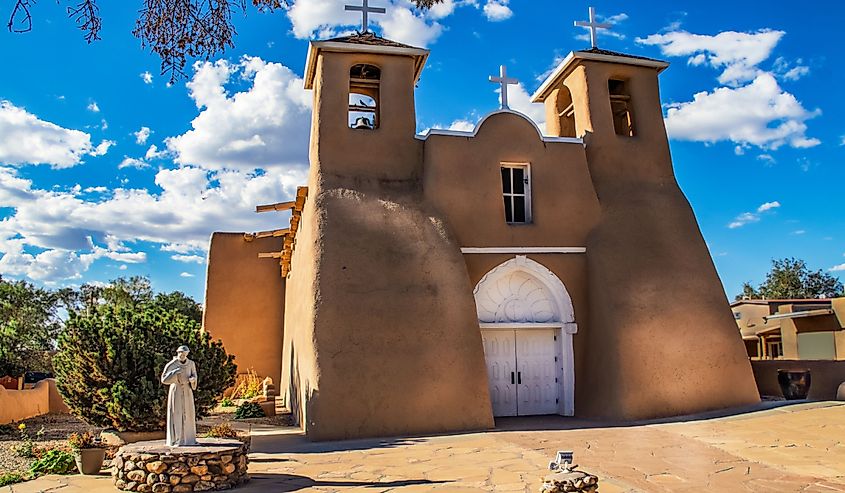 Historic adobe San Francisco de Asis Mission Church in Taos New Mexico in dramatic late afternoon light under intense blue sky with fluffy while clouds and birds in the belfry