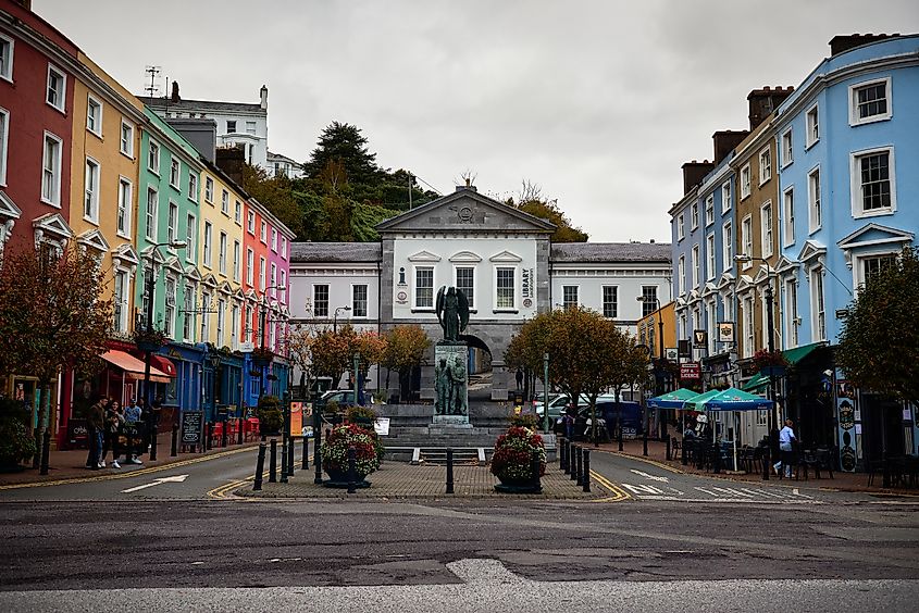 View of Casement Square, Cobh, featuring colorful Georgian buildings, the Lusitania Memorial, and vibrant street life.