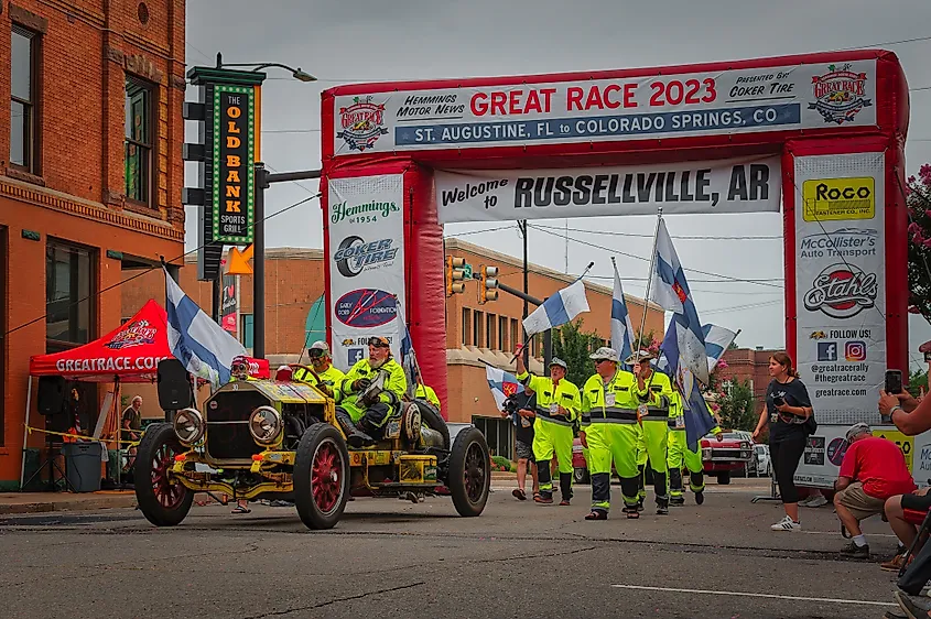 Vintage car under a red "Great Race 2023" arch in Russellville, AR. People in neon safety gear and flags, brick buildings in the background. Festive atmosphere.