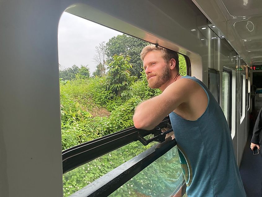 A blond man sticks his head out the window of a train to feel the breeze.
