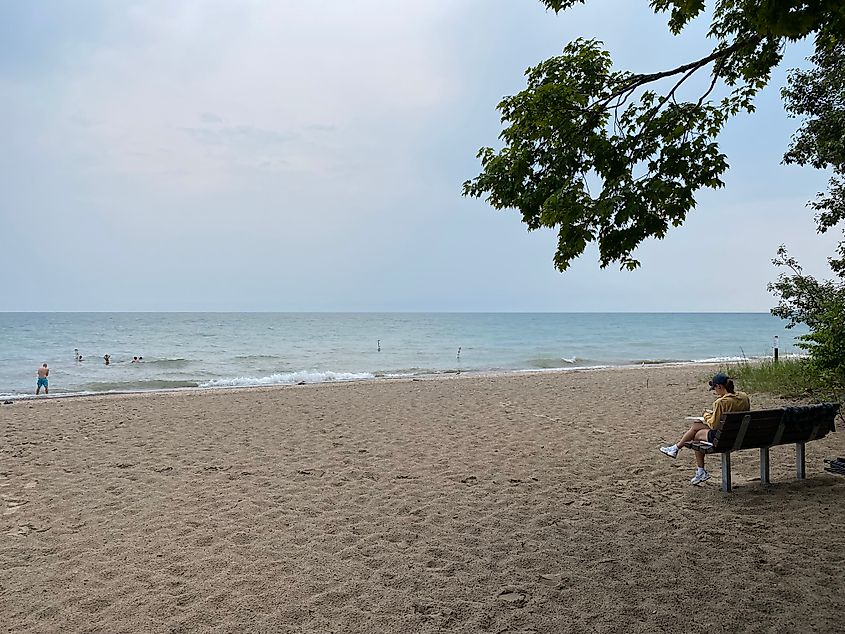 A woman eats lunch on a beachside bench while a family frolics in the water in the distance.