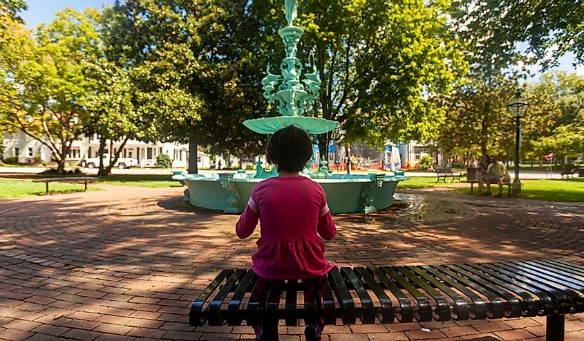 A little girl wearing pink casual clothing is sitting alone on a metal bench in the Fountain Park of Chestertown, Maryland