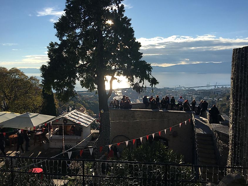 A crowd stands atop the lookout of a stone castle. A city and a bay can be seen far below.