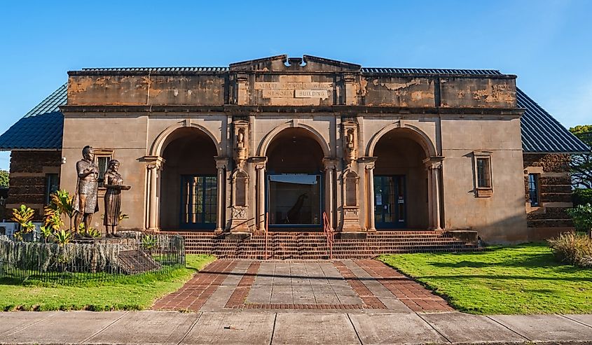 The Kauai Museum in Lihue, Hawaii features a classical facade with three arches, ornate columns, a statue, brick pathways, and a clear blue sky.