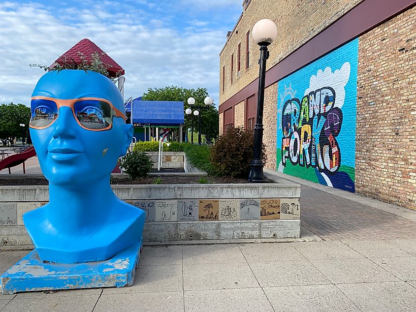 A large blue head sculpture wearing novelty sunglasses stands next to a colorful town mural for Grand Forks, North Dakota.
