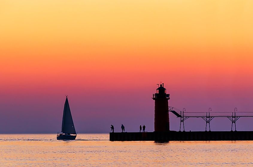 A vividly colorful twilight sky silhouettes a sailboat, people, and the lighthouse at South Haven, Michigan on Lake Michigan.