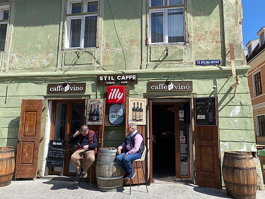 Two old men sit outside of a weathered cafe with whisky barrels standing in as tables. 