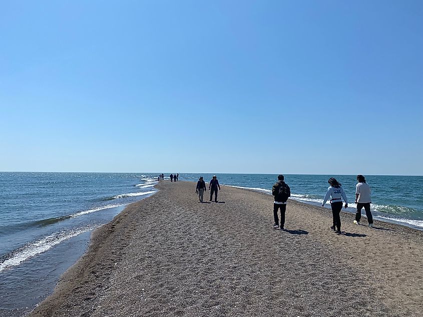 Small groups of people walk out to the tip of a narrow sand spit on a sunny day.