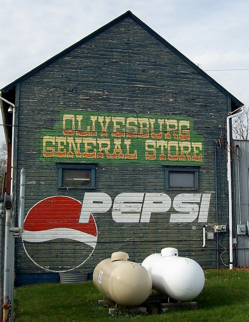 Weathered dark wooden side wall of a building painted with large vintage signs reading "Olivesburg General Store" and "Pepsi," with two propane tanks on the grass in front under a gray sky.
