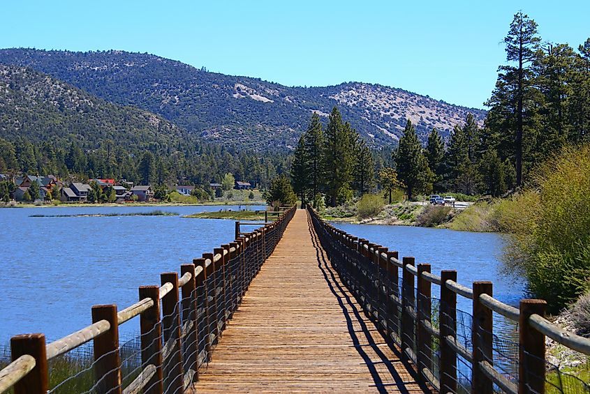 A wooden bridge spans across Big Bear Lake in California, offering a picturesque view of the serene waters surrounded by towering pine trees and rugged mountains.