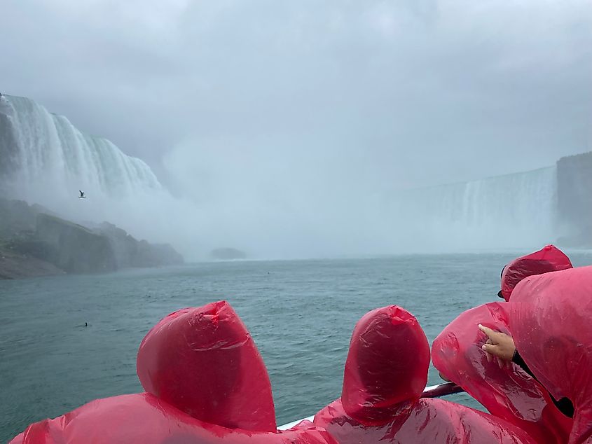 Passengers in red ponchos aboard a boat approaching a massive, curved, and misty waterfall on an overcast day.
