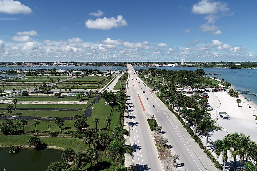 Aerial view of Fort Pierce Inlet in Florida with a bridge crossing the water, palm-lined roads, a sandy beach, and boats in the distance.