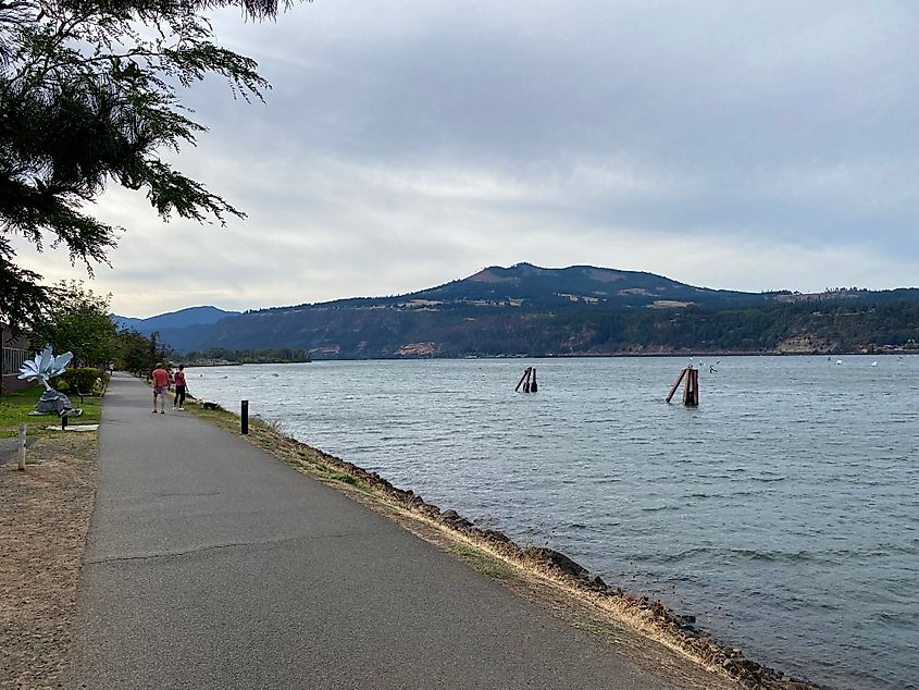 A paved waterfront path moves toward distant mountains. A group of windsurfers are faintly seen as well.