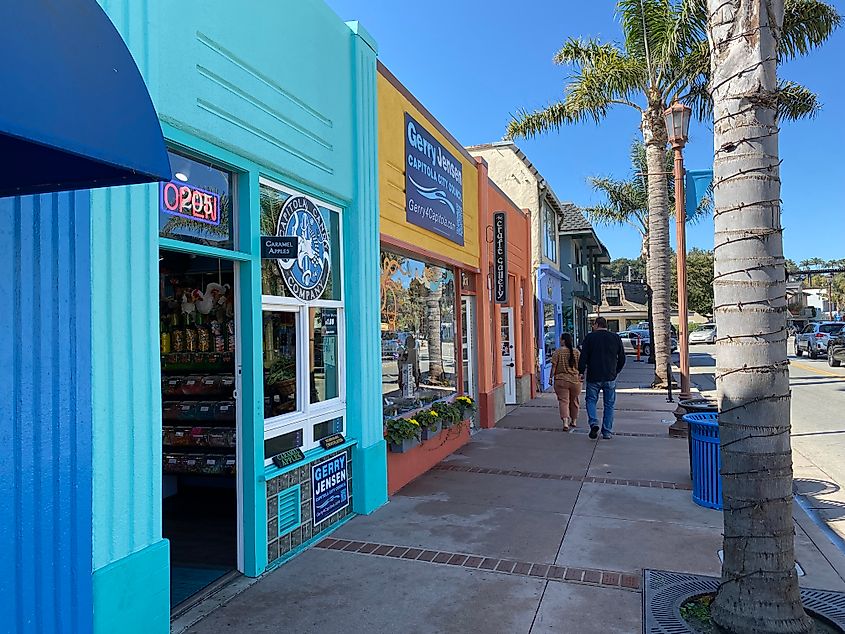 A couple walks along a palm-tree-lined sidewalk next to colorful boutique shops.