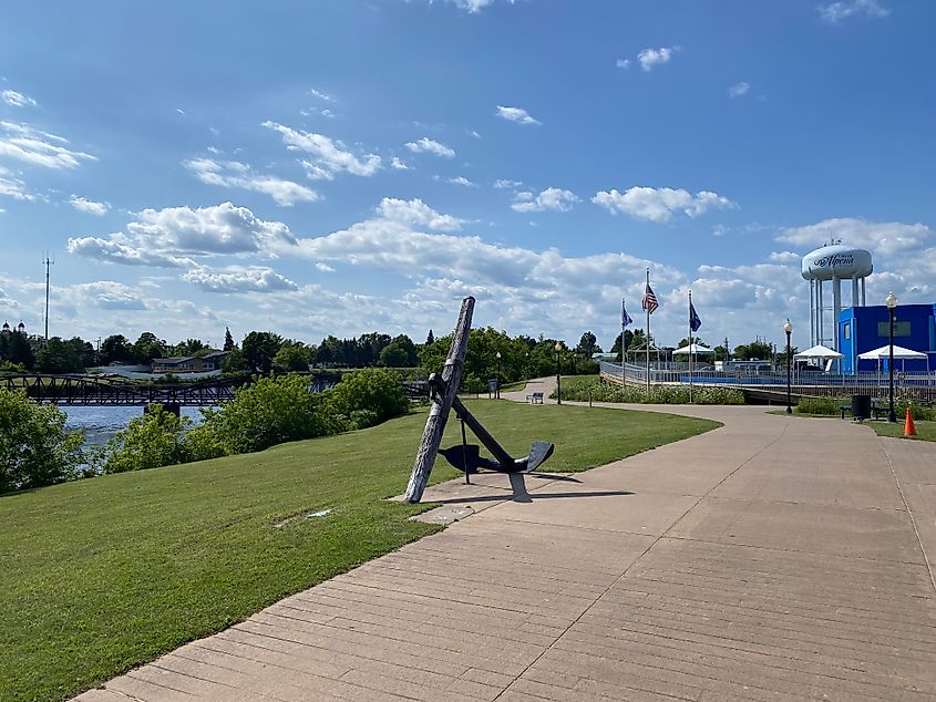 A large black anchor stands beside a riverside walking path. 