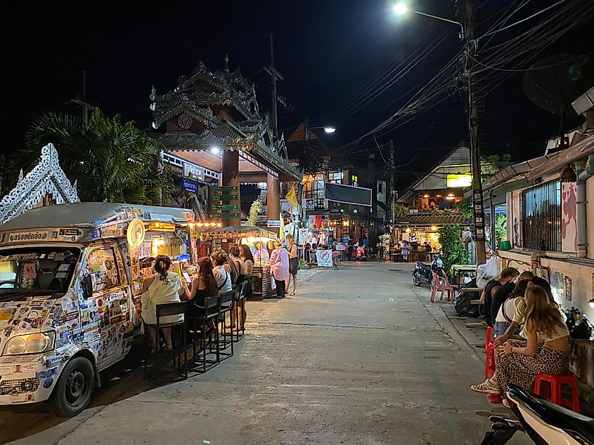 Youthful travelers congregate around a sticker covered food truck at night on a slim street in Thailand.