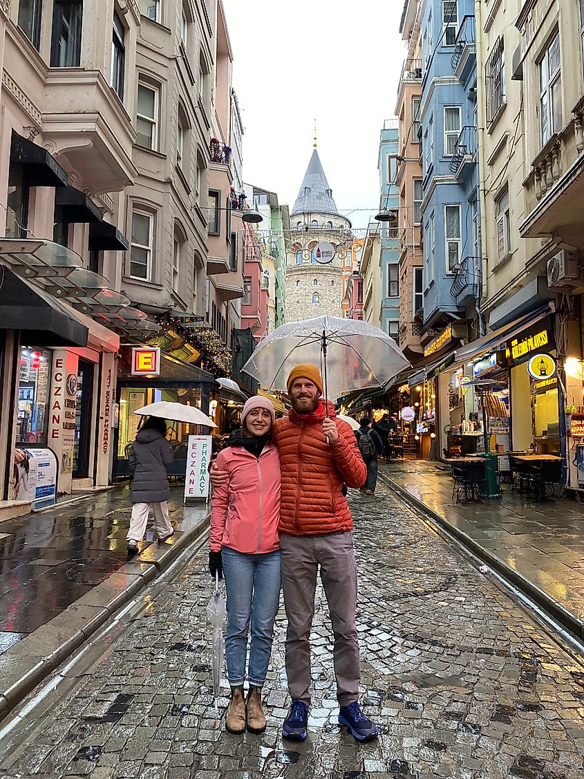 A woman in a pink jacket and a man in an orange one huddle under an umbrella, posing for a picture in front of the medieval Galata Tower.
