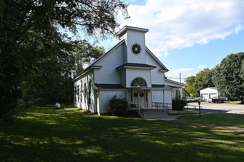 A quaint white church with a steeple and a round window, surrounded by lush greenery under a clear blue sky, in Bell Buckle, Tennessee, during the fall season.