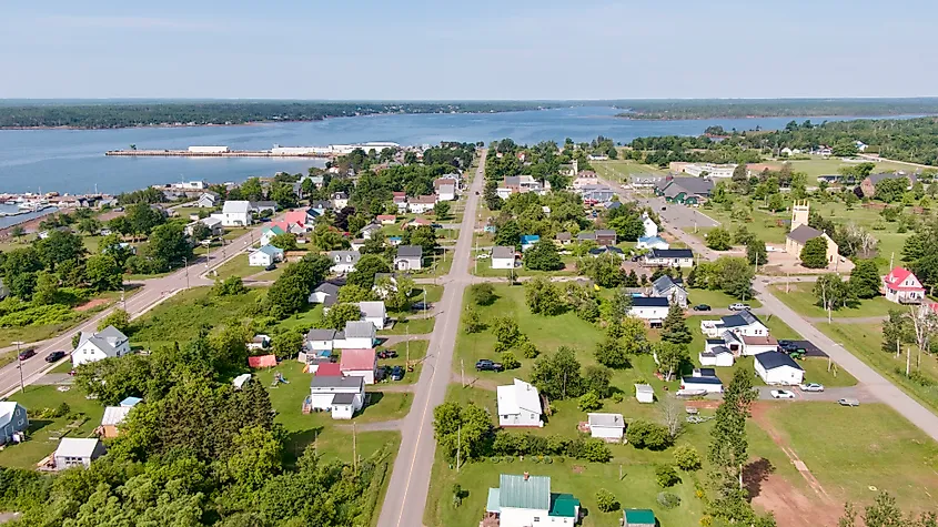 Aerial view of a small coastal town with tree-lined streets and charming houses, bordered by a calm blue waterway under a clear sky, conveying tranquility.