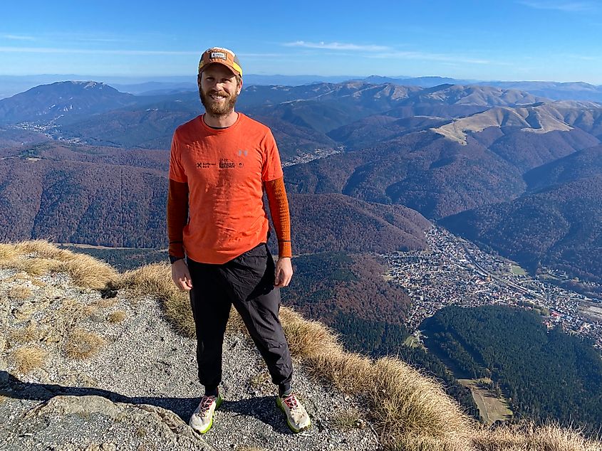 A man in athletic gear poses for a cliffside picture, high above a mountain town seen below