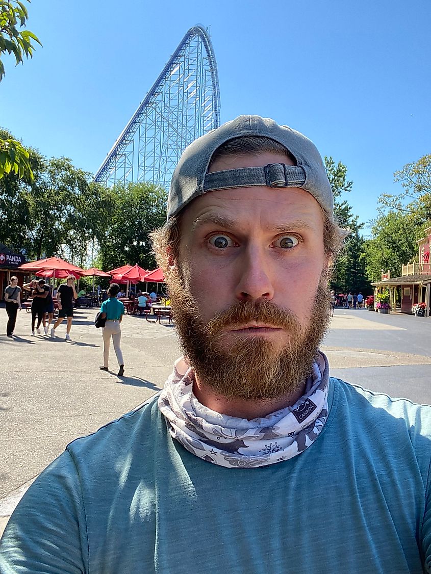 A man makes a "gonna puke" face while taking a selfie in front of a massive roller coaster. 