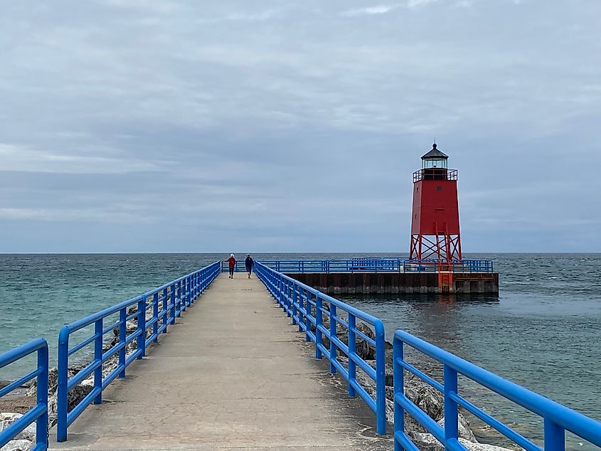 Two women walk a lakeside pier toward a red lighthouse on an overcast day.