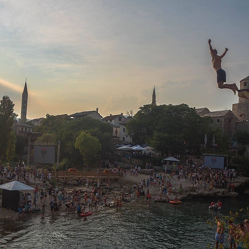 A man jumps off a high platform into a river while crowds from the recent Red Bull event begin to disperse into Mostar's Old Town