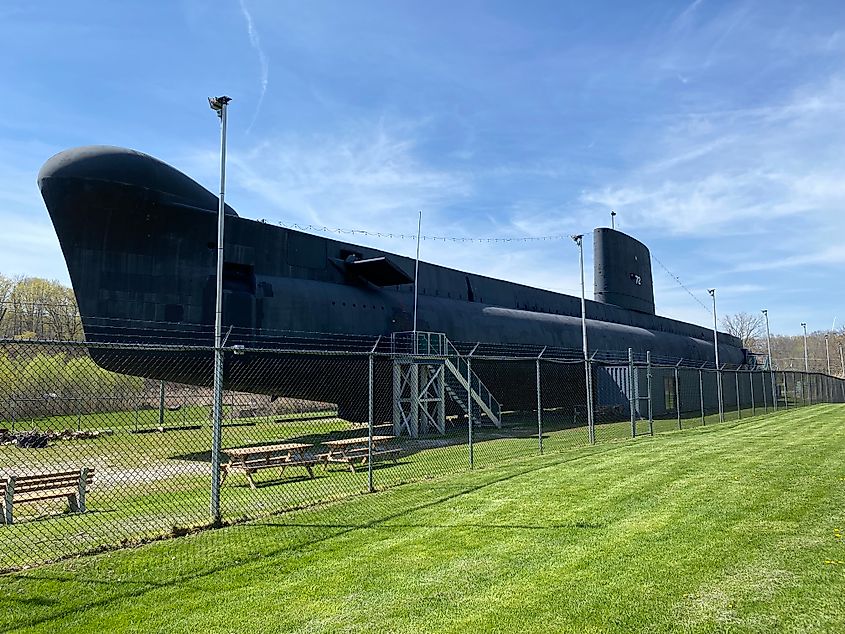 A large black submarine sits inside of a chain link fence in a large grassy field.