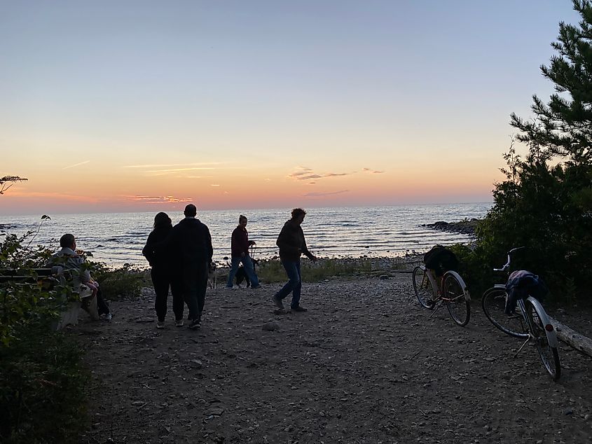 Campers silhouetted by a pastel sunset while standing on a small rocky beach.