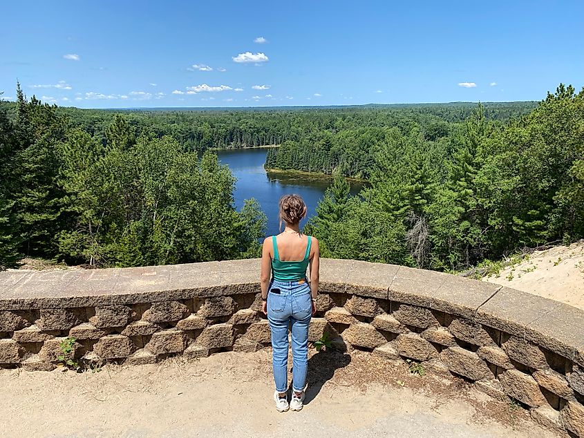 A woman in jeans and a green tank top looks out over a forested river from atop a sand dune lookout. 