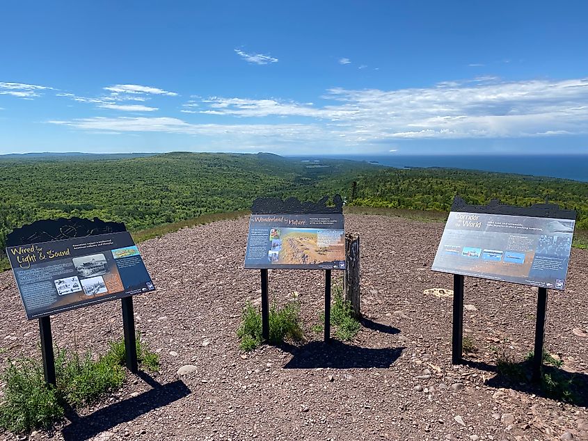 Interpretive signs line a mountain top viewpoint. Sprawling evergreens and the blue of Lake Superior occupy the panorama. 