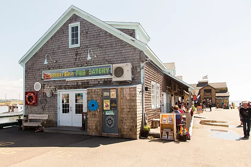 Rustic coastal pub and eatery with weathered wood siding, colorful signage, patrons dining outside, and a sunny, relaxed atmosphere.