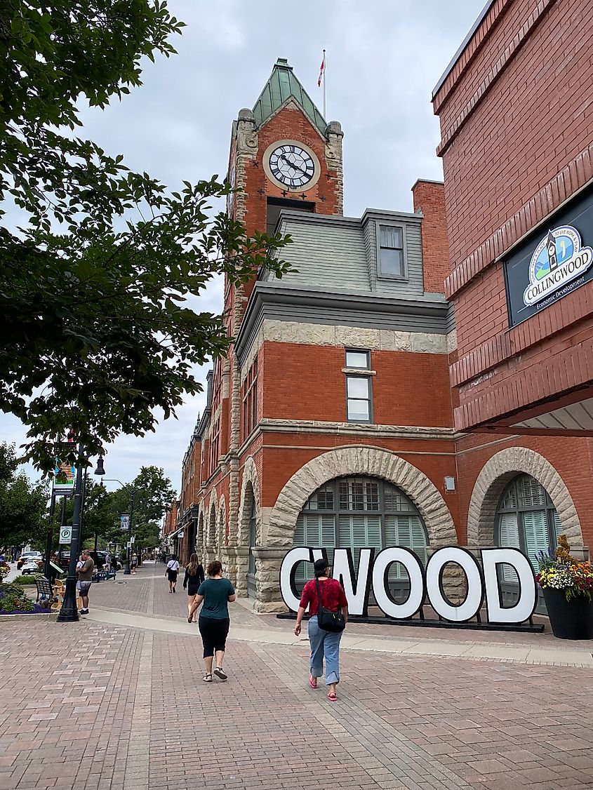 Pedestrians walking past a large, red-brick clock tower and white block letters spelling "CWOOD" for Collingwood.