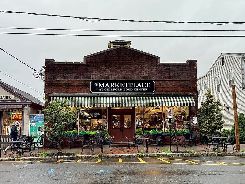Front view of The Marketplace at Guilford Food Center in Guilford, Connecticut, with outdoor seating, striped awning, and people inside the café.