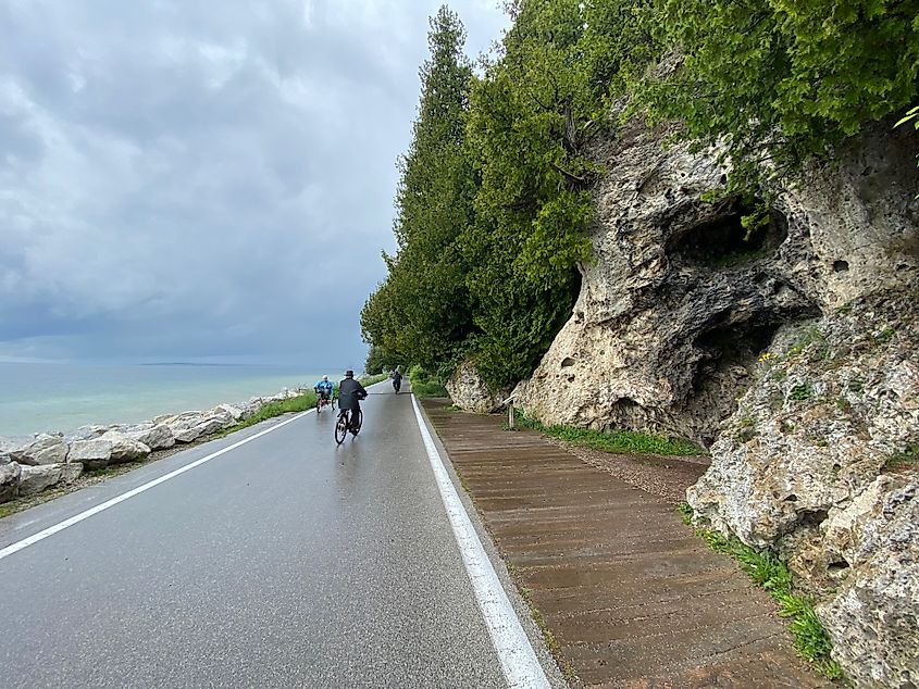 A few cyclists pedal around a lakeside bike path walled on the right by karst, tree-lined cliffs