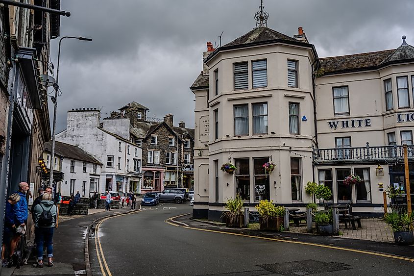 Pedestrians walk along a sidewalk near the White Lion Hotel in the town of Ambleside. Traditional slate and stone architecture lines the curved road under an overcast sky. Ambleside, United Kingdom.