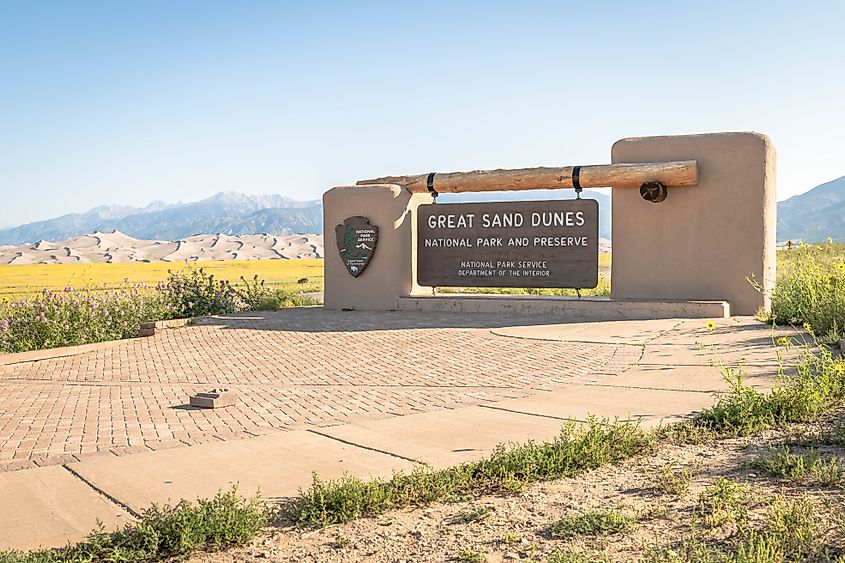 Editorial Photo Credit: Keith J Finks via Shutterstock. ALAMOSA, CO - 14 AUGUST 2021: The sign at the entry to Great Sand Dunes National Park on a crystal clear summer day.