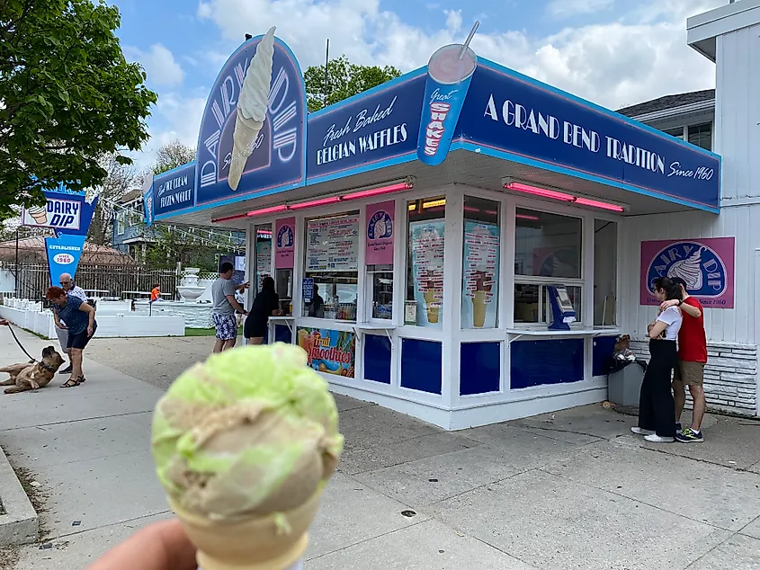 An ice cream cone held up in front of an old fashioned ice cream parlour in the middle of a summer beach town.